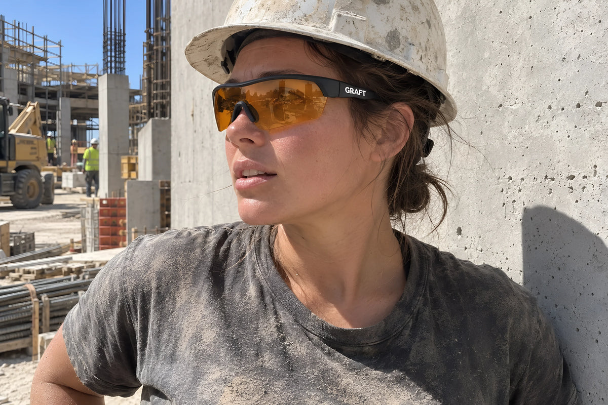 A woman in a dusty hard hat and a dust-covered work t-shirt wearing the amber GRAFT Amber glasses, standing against a concrete wall taking a breather, looking off to one side. Daytime, hard sun, no candles lit.