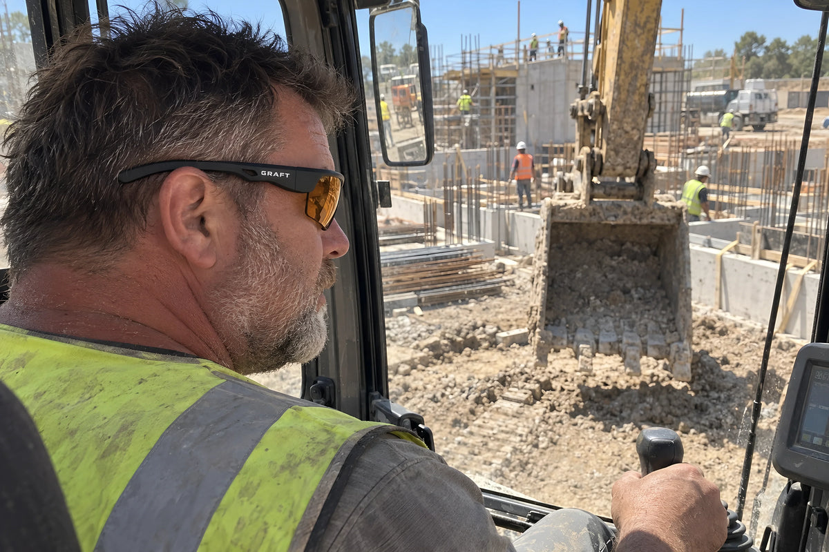A plant operator in the cab of a digger wearing amber GRAFT glasses, one hand on the joystick, looking out over the bucket. Daytime, hard sun, no candles lit.