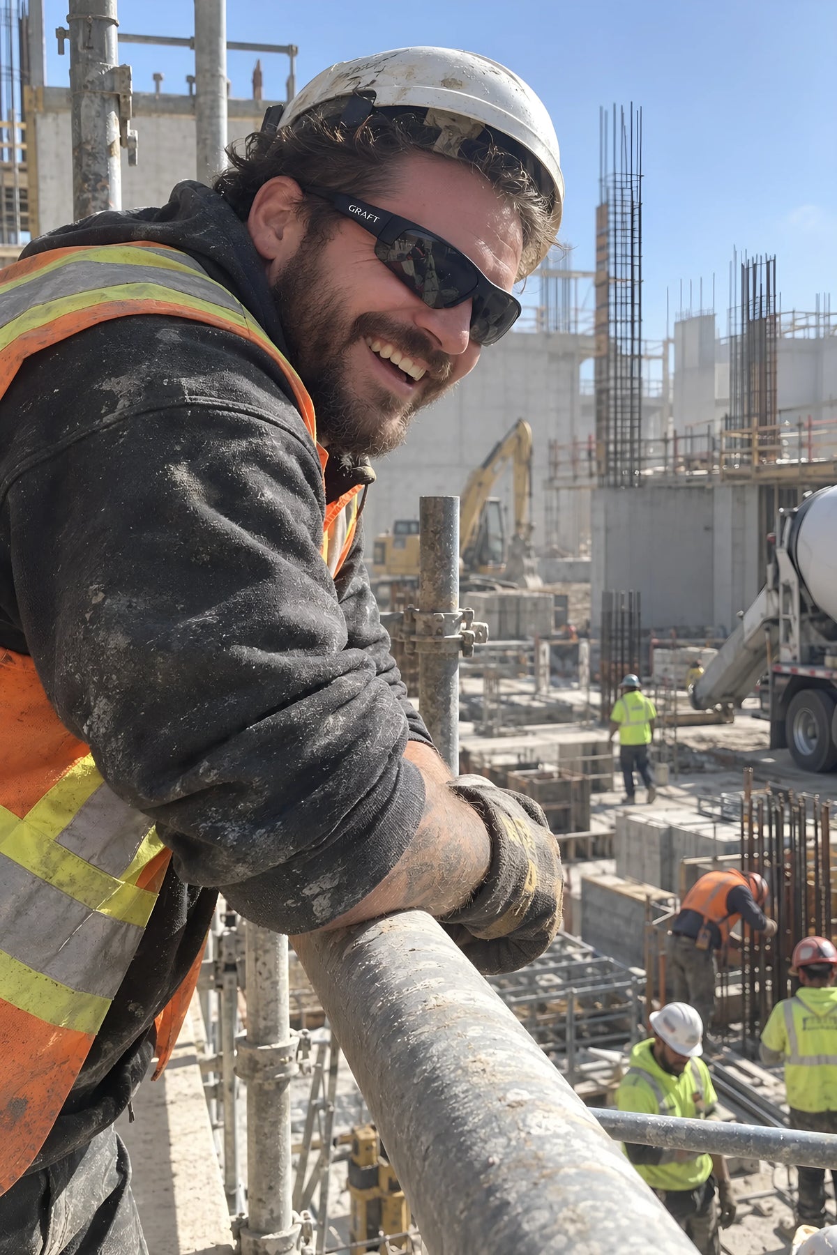 A scaffolder leaning on a guard rail high up in smoke GRAFT glasses, hard hat pushed back, grinning down at whoever is below. Daytime, hard sun, no candles lit.