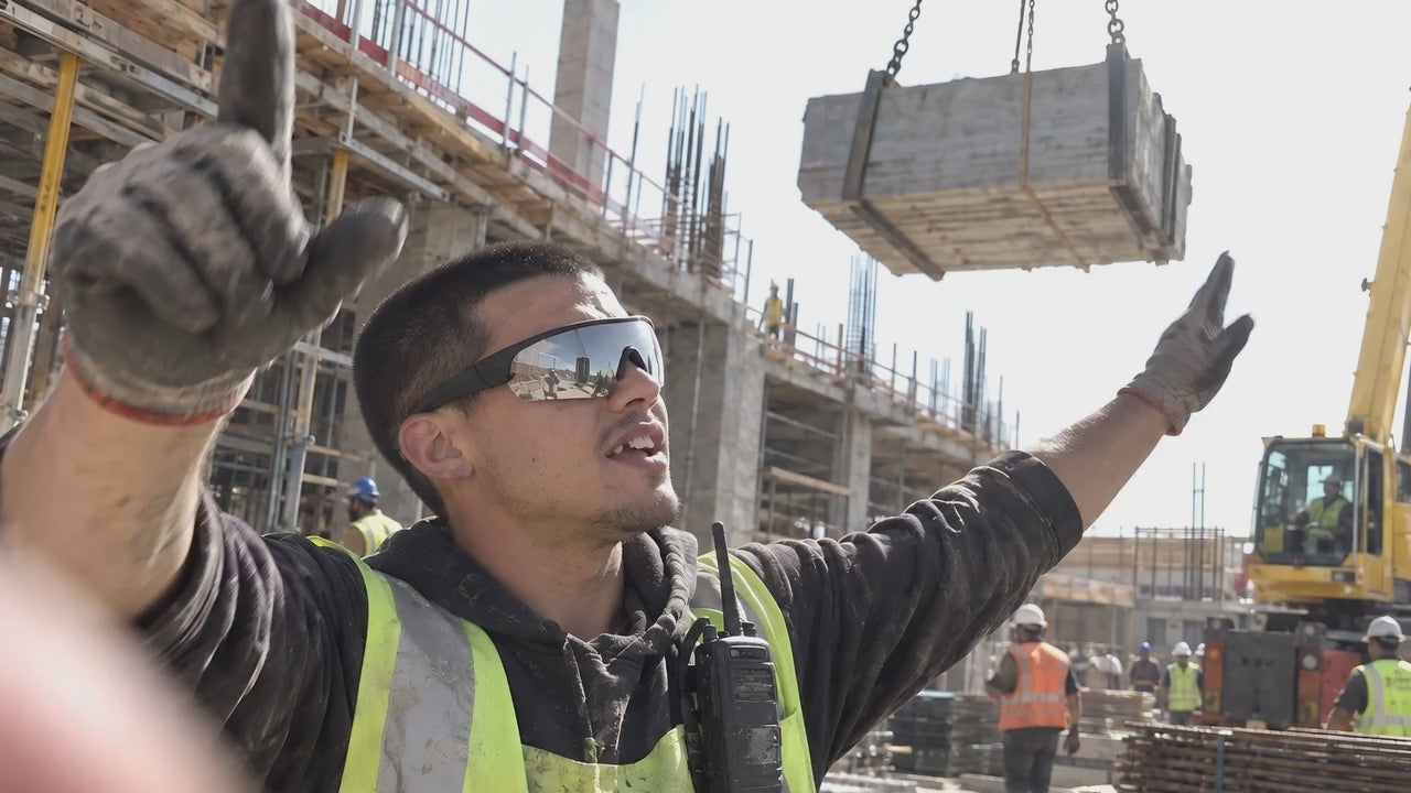 A banksman in mirrored GRAFT glasses signaling a crane, the load swaying above him